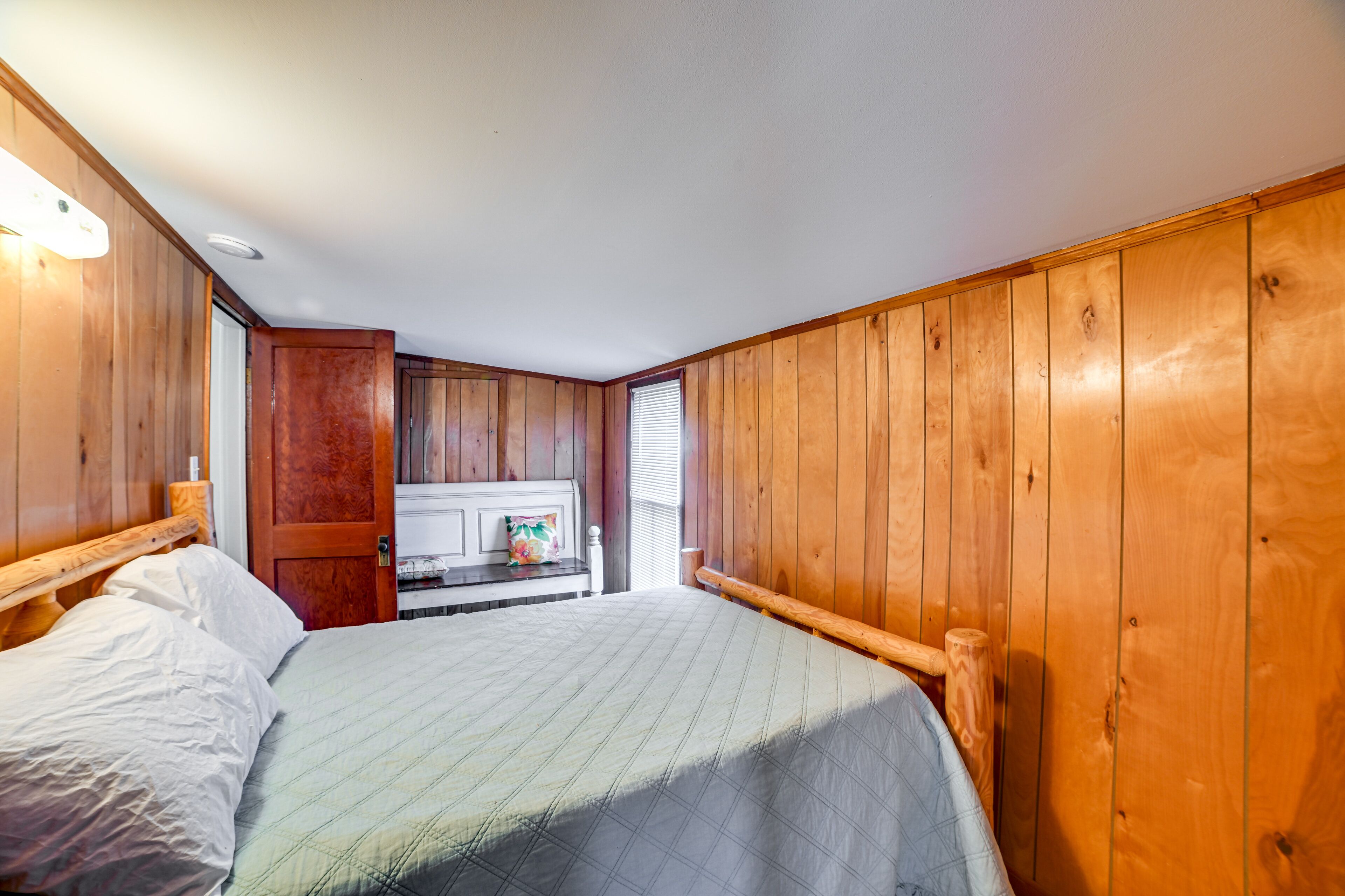 Ein Schlafzimmer mit Holzwänden, einem Bett mit weißer Bettwäsche, einem Holz-Kopfteil und einer kleinen Küchenzeile im Hintergrund.