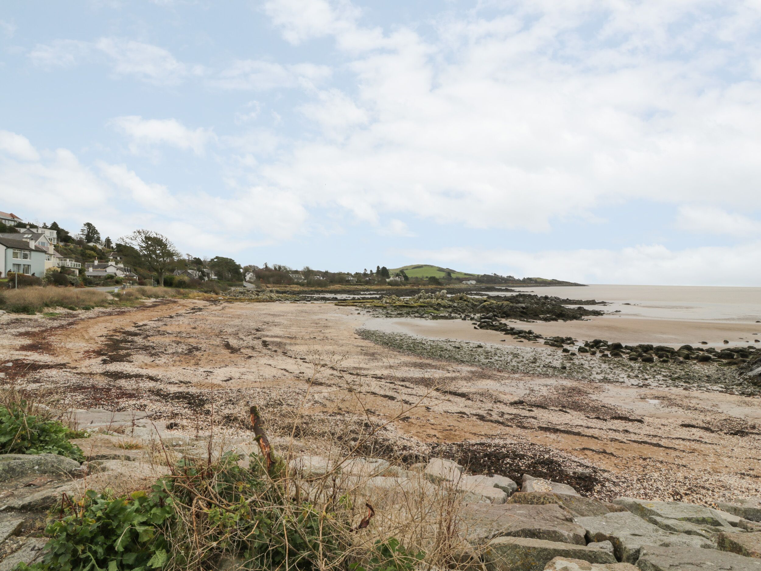 A coastal landscape with a sandy beach, rocky outcrops, and a distant view of houses on a hill.
