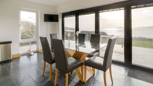 A modern dining area with a glass table and chairs, a flat-screen TV, and a view of the sea. - Merse End (Castle Douglas)