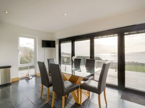 A modern dining area with a glass table and chairs, a flat-screen TV, and a view of the sea.