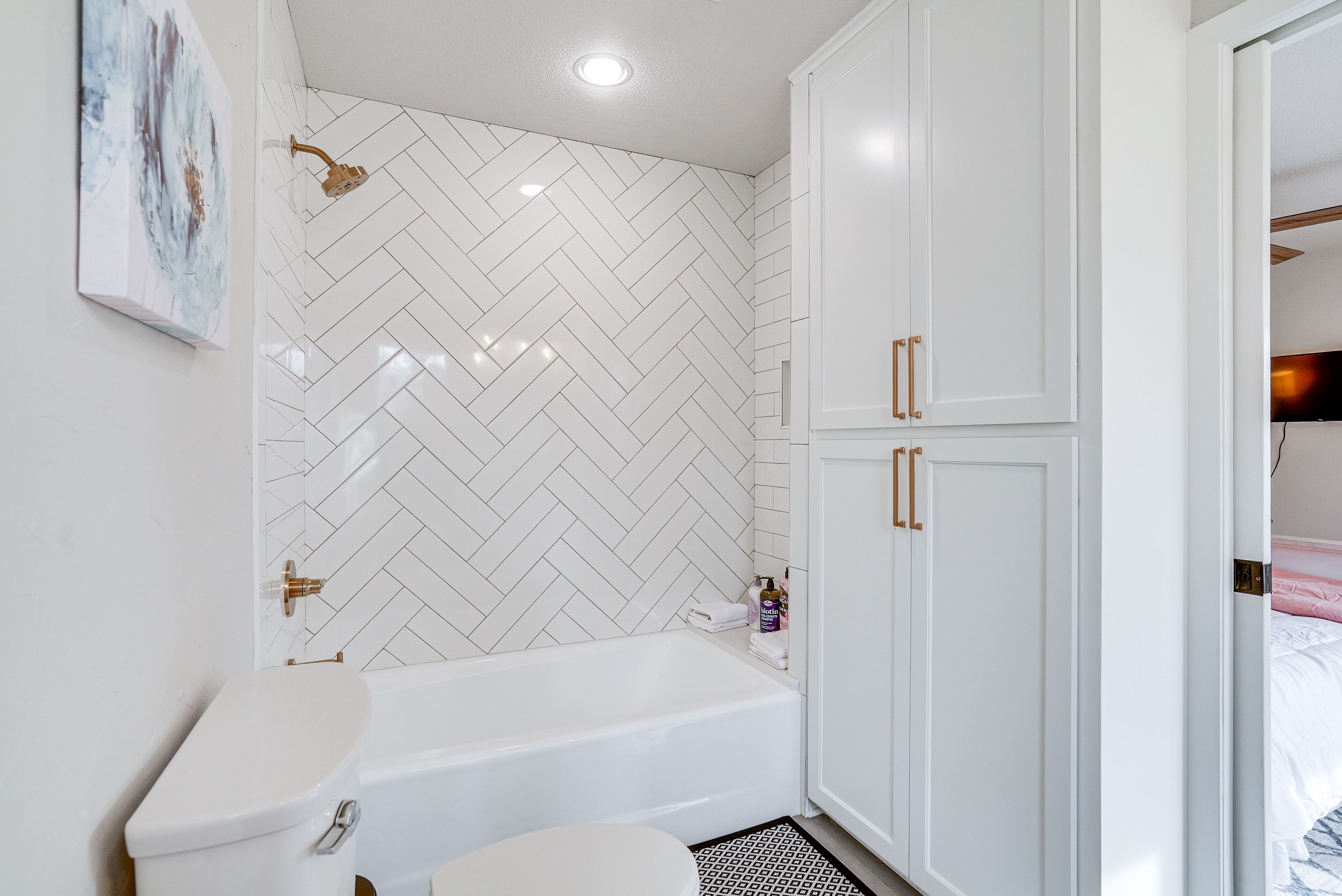 A bathroom with a white herringbone tile backsplash, a white bathtub, and a toilet. There are white cabinets with gold handles and a small shelf with toiletries.