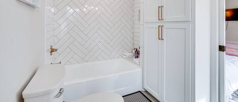 A bathroom with a white herringbone tile backsplash, a white bathtub, and a toilet. There are white cabinets with gold handles and a small shelf with toiletries.