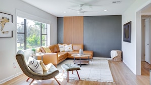 Un salon moderne avec un canapé d’angle, une table basse ronde et un fauteuil. On y trouve un ventilateur de plafond, une fenêtre donnant sur un espace verdoyant et une étagère fixée au mur.