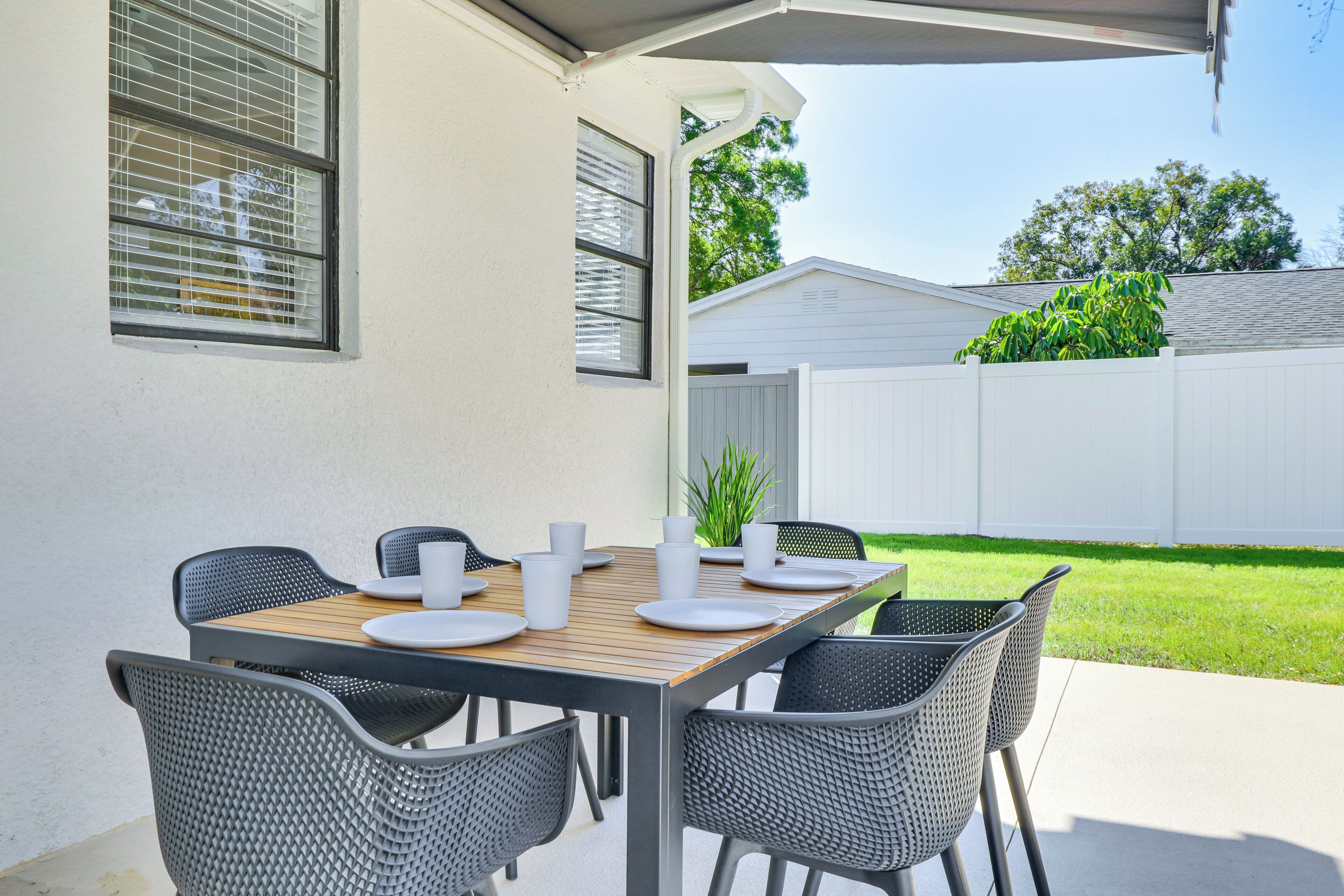 A patio with a table set for four, surrounded by wicker chairs, against a white fence and a green lawn.