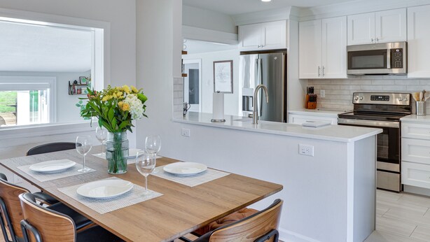 Une cuisine moderne avec des meubles blancs, des appareils en acier inoxydable et une table à manger en bois pour quatre personnes.