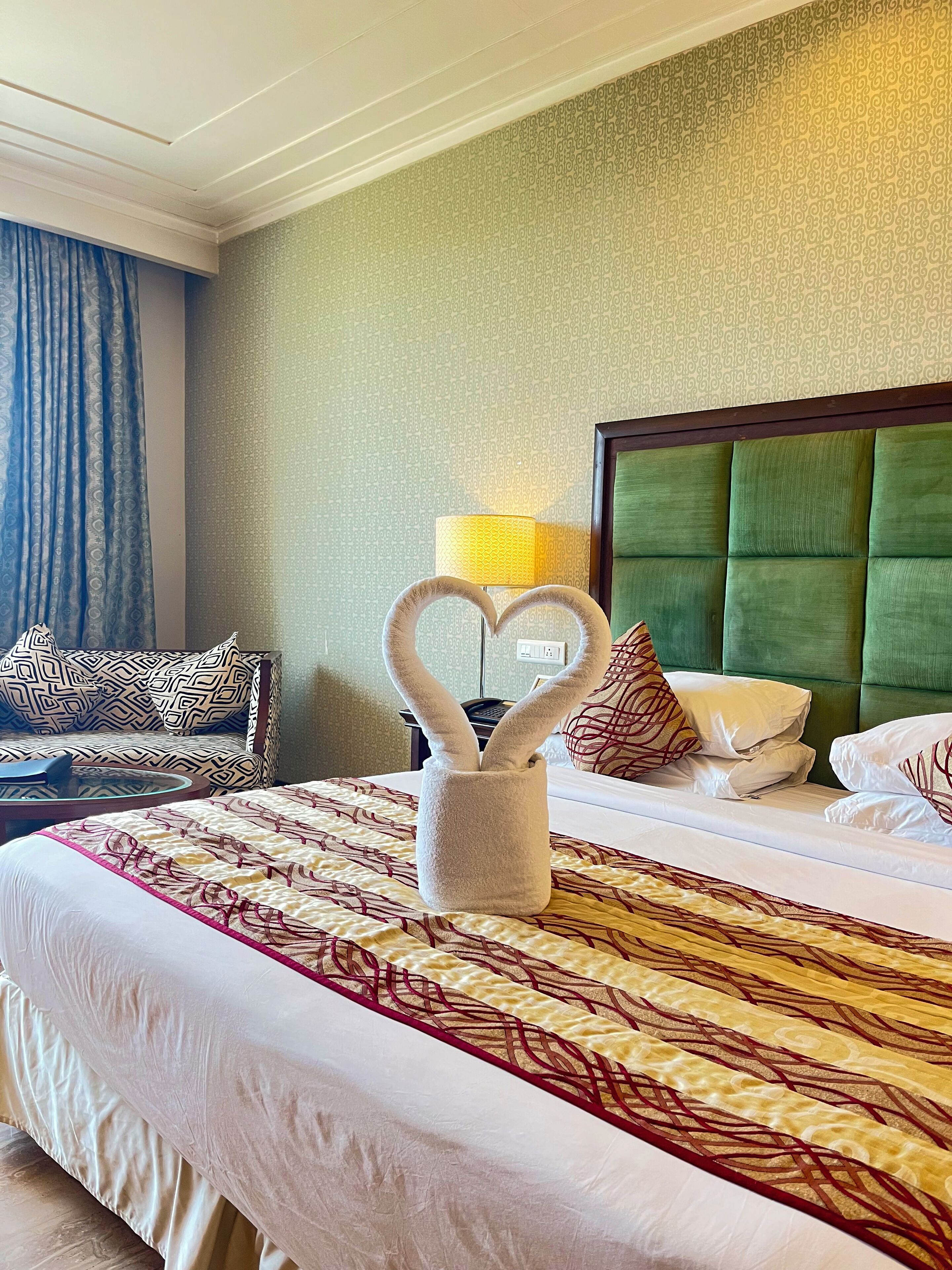 A hotel room with a large bed, a heart-shaped towel holder, a green upholstered headboard, and patterned bedding.