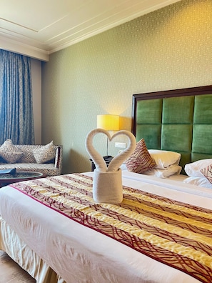 A hotel room with a large bed, a heart-shaped towel holder, a green upholstered headboard, and patterned bedding.