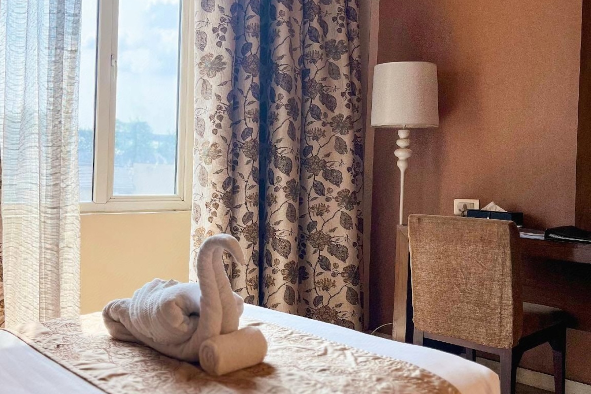 A hotel room with a bed, a desk, a chair, a lamp, and a window with curtains.