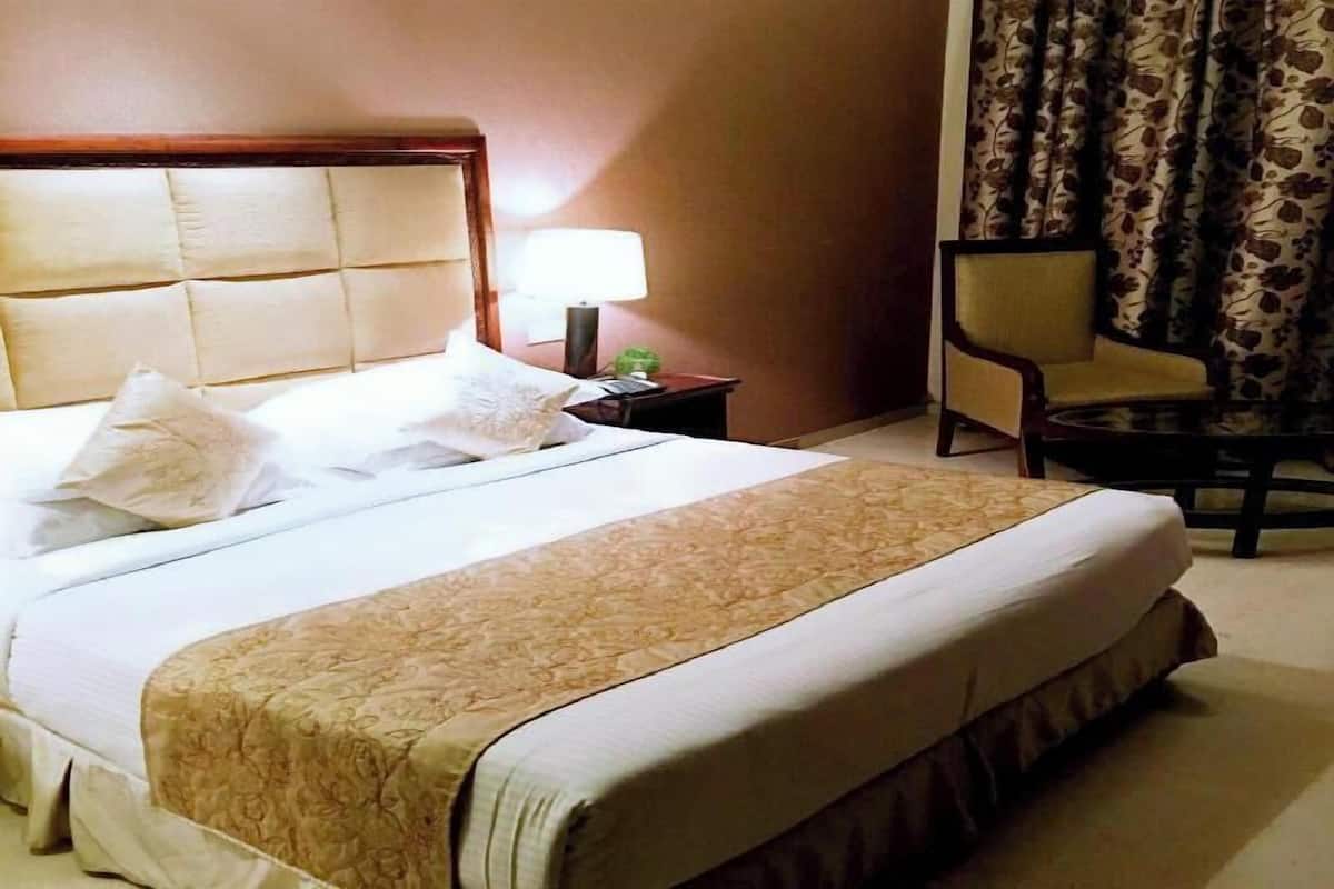 Quarto de hotel com uma cama grande, mesa de cabeceira com abajur, uma cadeira e vista para o exterior através das cortinas.