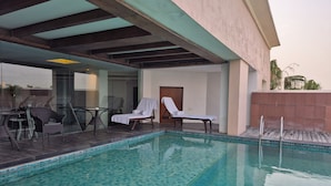 A swimming pool with lounge chairs and a table, surrounded by a tiled deck and a glass wall. - Godwin Meerut (Meerut)