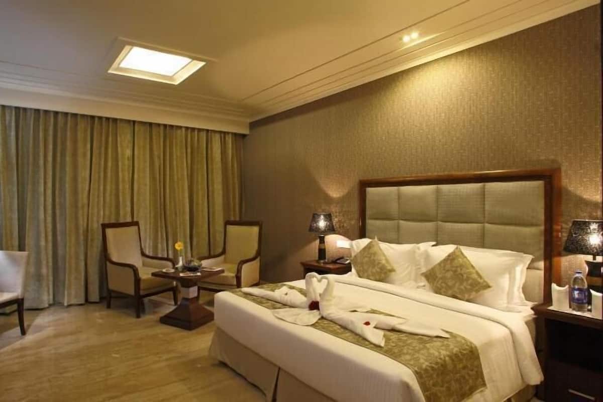 Quarto de hotel com cama, duas cadeiras, escrivaninha e uma janela grande com cortinas.