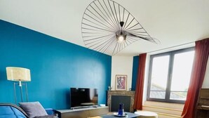 A modern living room with a blue sofa, a flat-screen TV, a fireplace, and a large window with curtains.