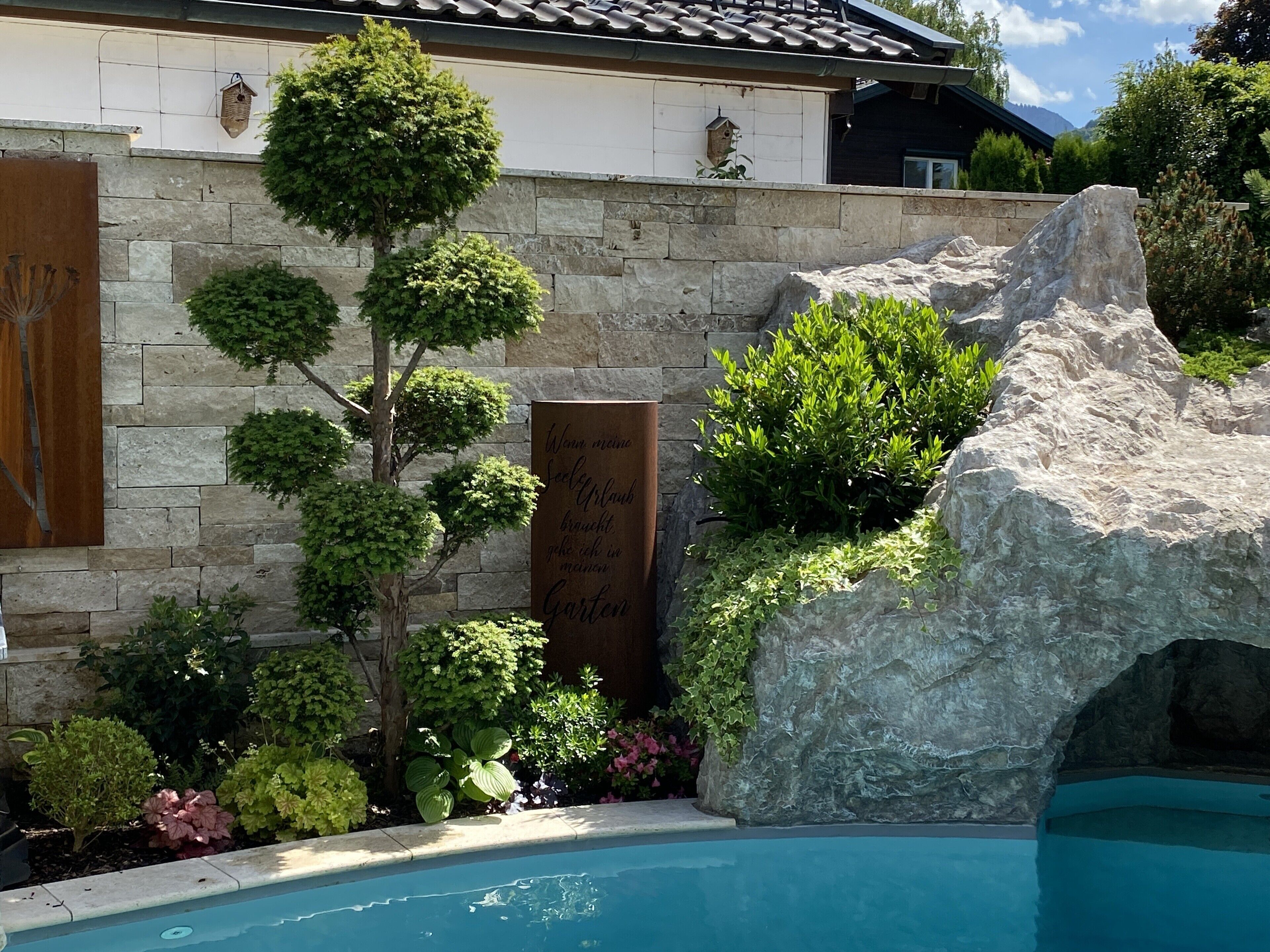 Eine Steinsäule mit einer Gedenktafel, ein kleiner Baum und eine Felsformation in der Nähe eines Pools.