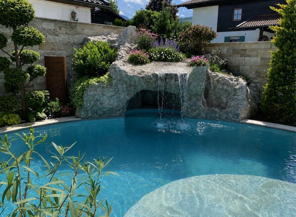 A swimming pool with a waterfall feature, surrounded by stone walls and lush greenery.