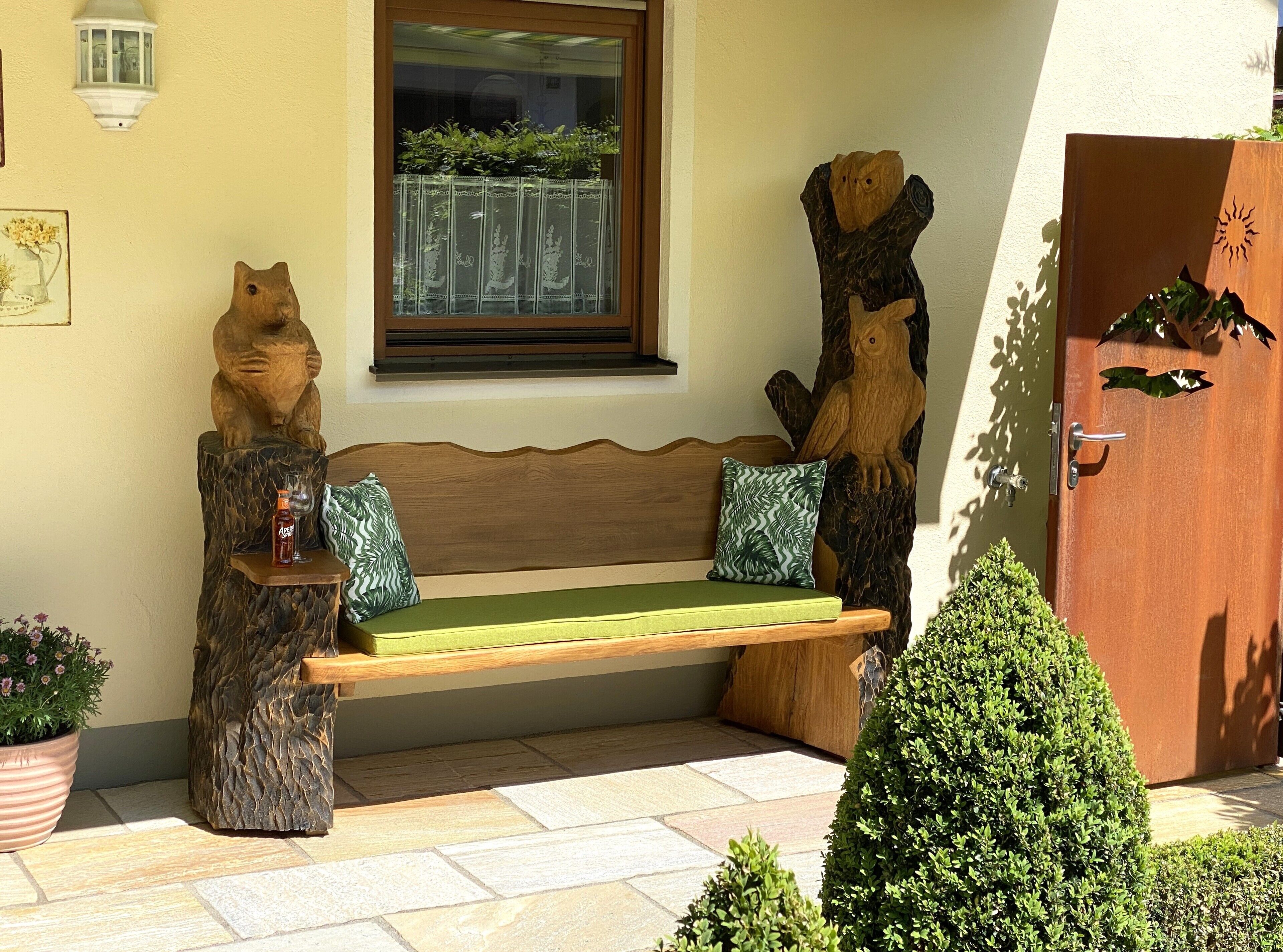 A wooden bench with a green cushion, two wooden animal sculptures, a potted plant, and a door with a sun cut-out design.