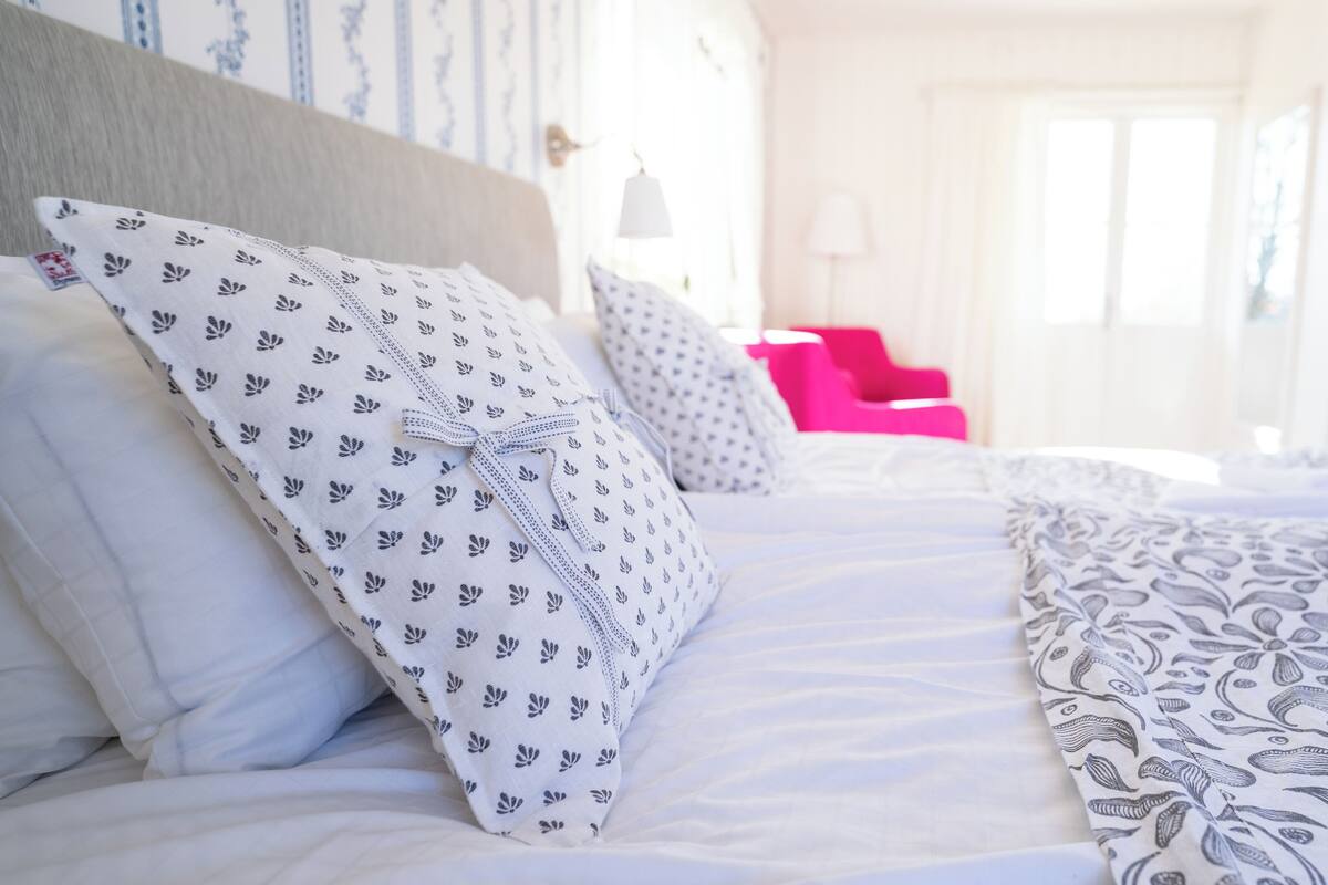 A neatly made bed with patterned pillows and a blanket, a pink chair, and a door leading to another room.