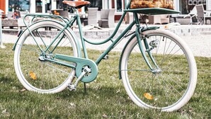 Una bicicleta color turquesa con una canasta que contiene pan y una botella, estacionada sobre césped frente a un edificio de estilo granero rojo.