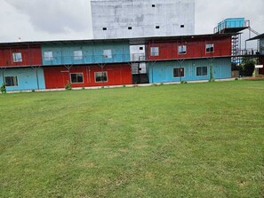 A building with multiple red and blue sections, a grassy field in front, and a large grey structure in the background. - Vrindavan Resort (Mathura)