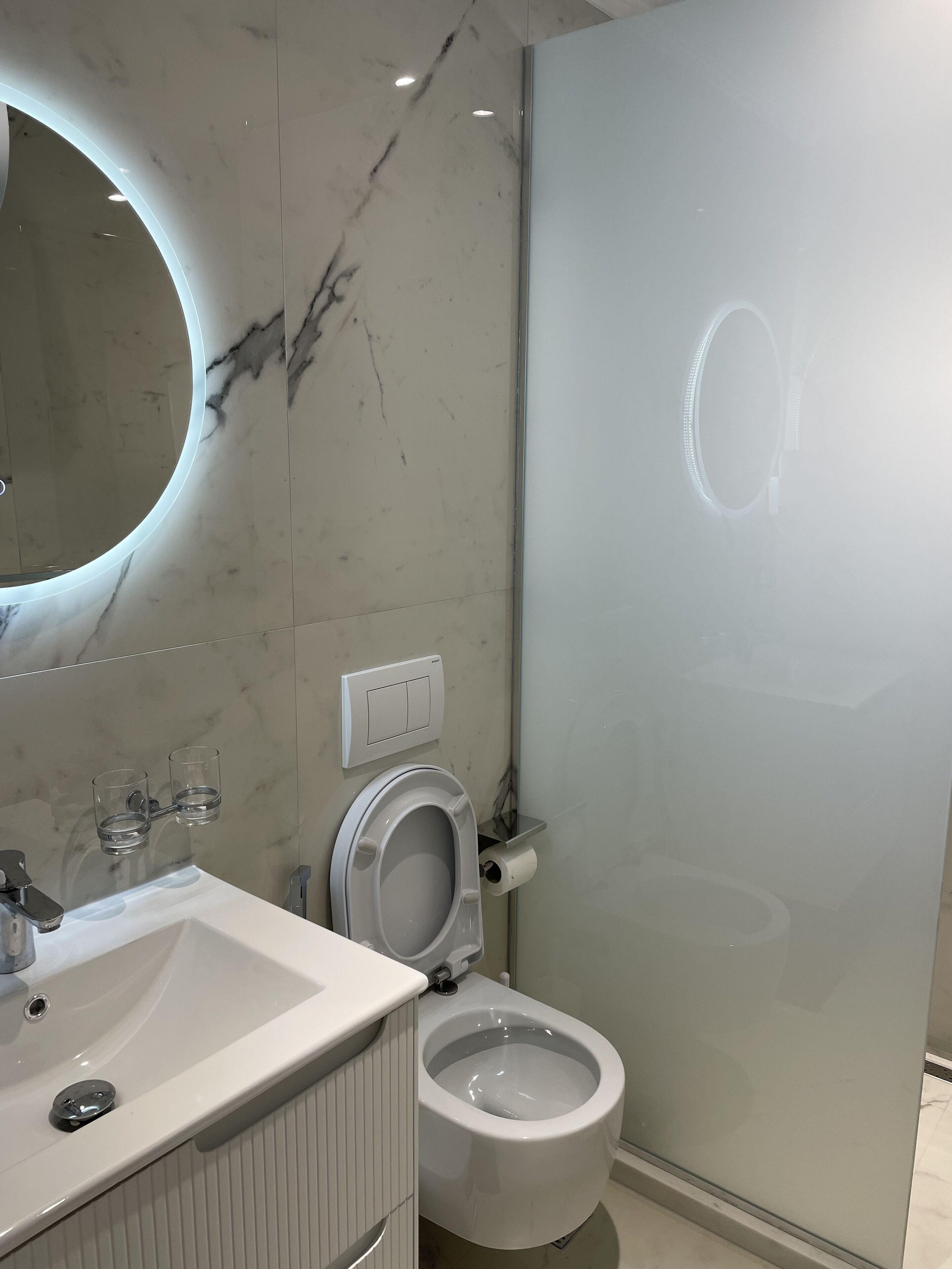A modern bathroom with a marble backsplash, a round mirror, a toilet, and a glass shower enclosure.