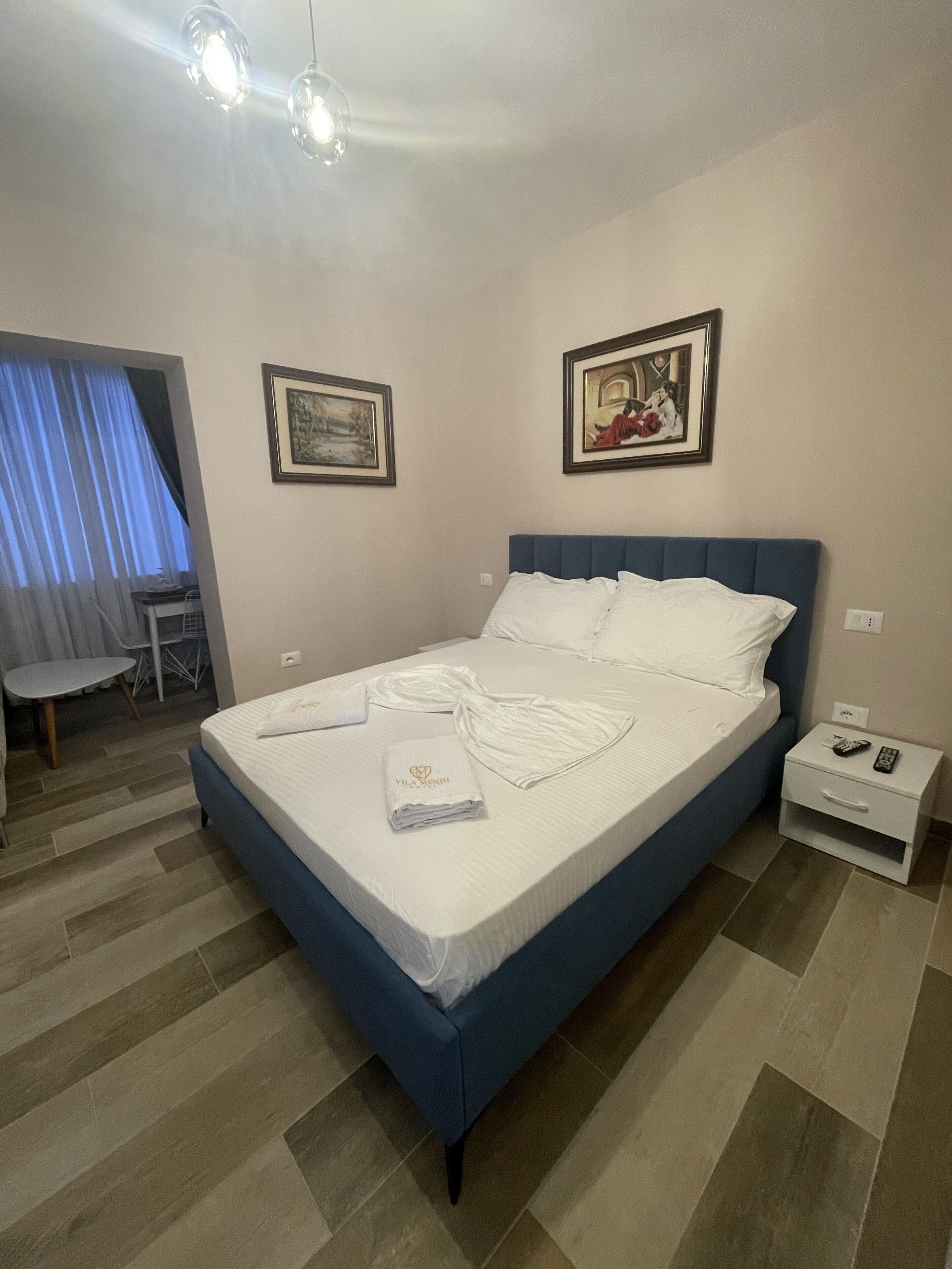 Ein ordentlich gemachtes Bett mit weißen Leinen und einem gefalteten Handtuch, ein Nachttisch mit Fernbedienung und zwei gerahmte Bilder an der Wand.