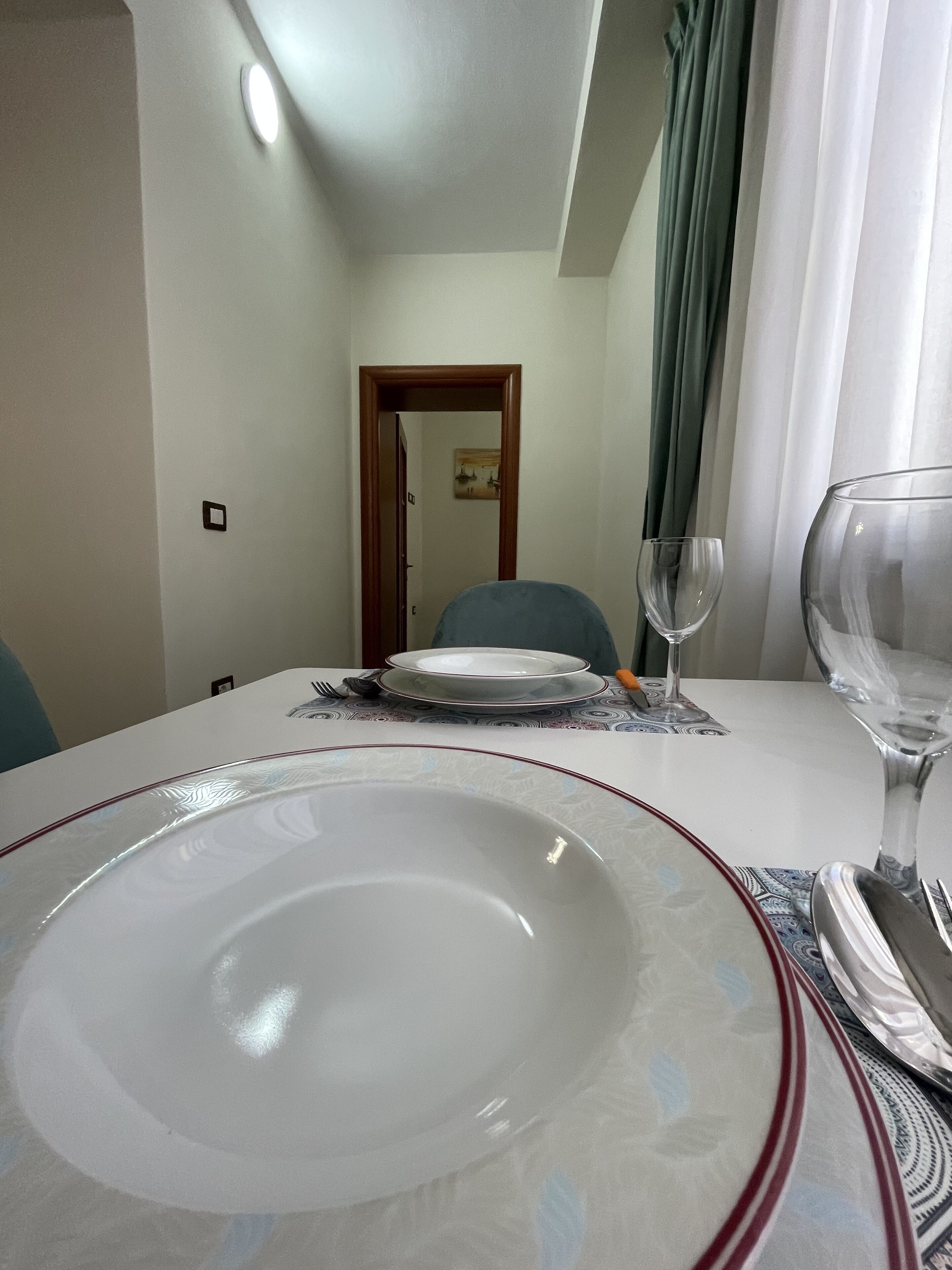 A dining table set with plates, cutlery, and a wine glass, with a view of a corridor and a door in the background.