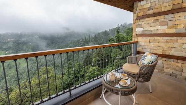 Un balcón con vistas a un valle boscoso, una silla de mimbre, una pequeña mesa redonda con una tetera y tazas, y un muro de ladrillo.