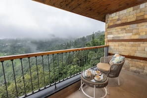 A balcony with a view of a forested valley, a wicker chair, a small round table with a teapot and cups, and a brick wall. - juSTa Hollow Oak Mussoorie (Dehradun)