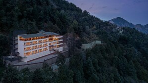 Un hotel moderno de varios pisos con balcones, ubicado en una ladera con un bosque denso y montañas al fondo.