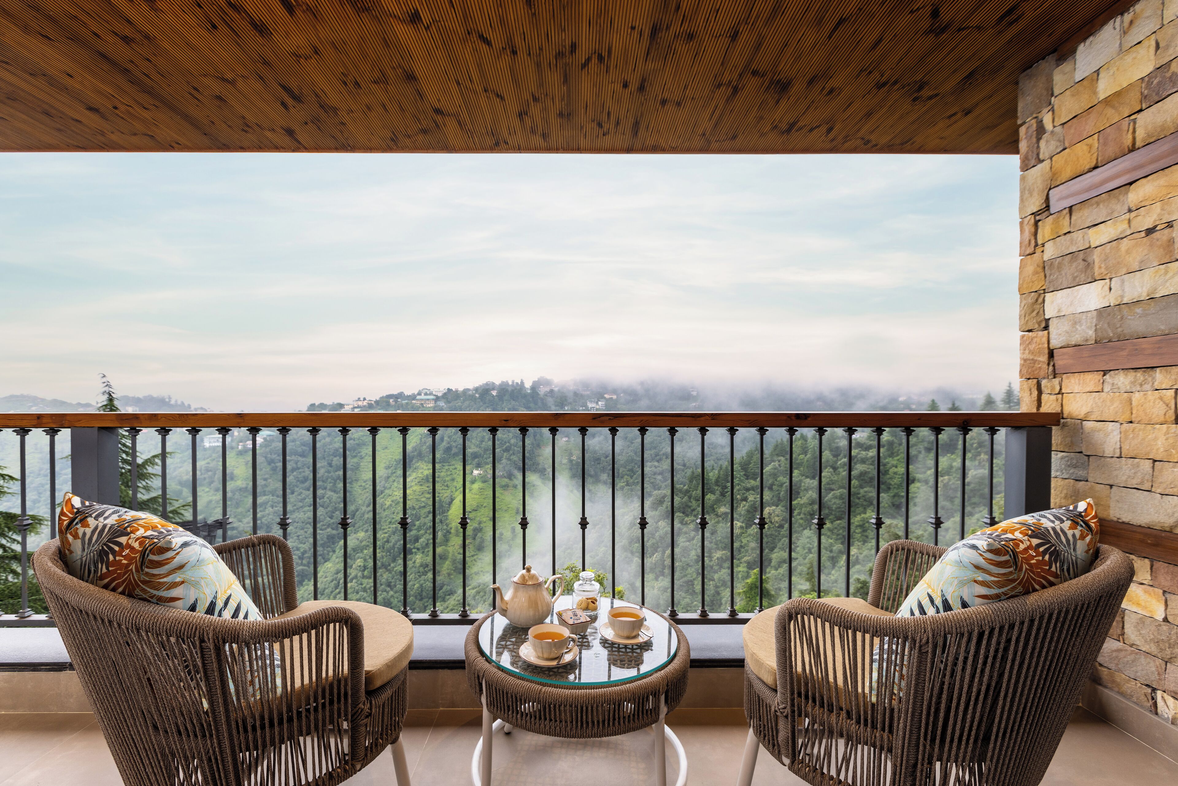 A balcony with wicker chairs, a table with tea set, and a view of a mountainous landscape.
