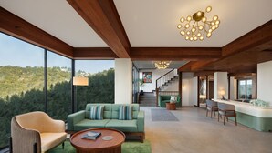 Amplia sala de estar con un gran ventanal que ofrece vistas a una ladera boscosa, un sofá verde, una mesa de centro de madera y una escalera que conduce a un nivel superior.