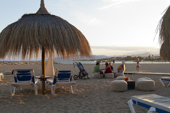 A beach with lounge chairs, a thatched umbrella, and a stroller.