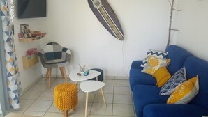 A living room with a blue sofa, a small white table, a yellow ottoman, a chair, and a surfboard on the wall. - Appartement Village de Vacances (Parentis-en-Born)
