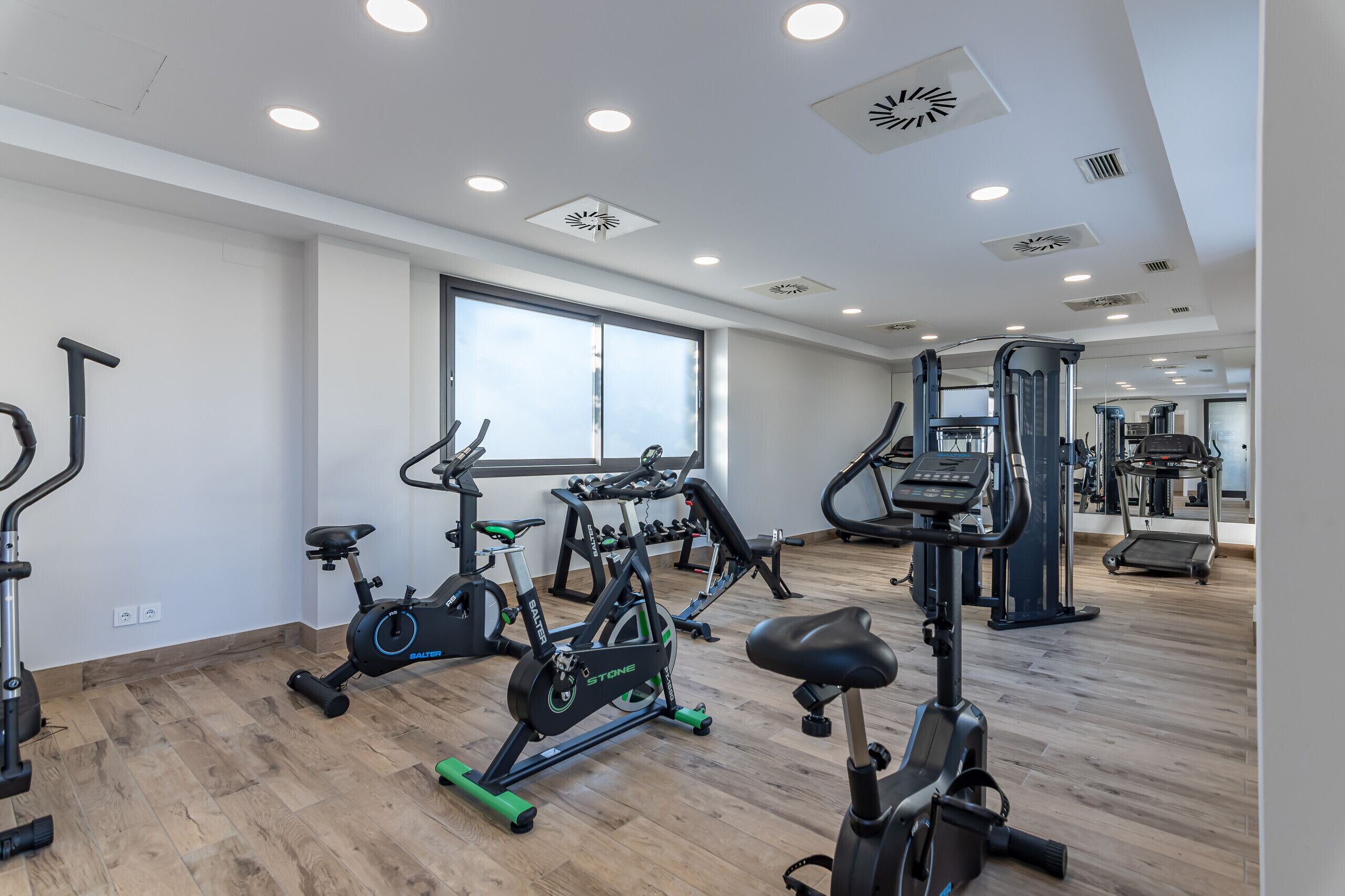 Een sportschool met diverse fitnessapparaten, grote ramen en een houten vloer.