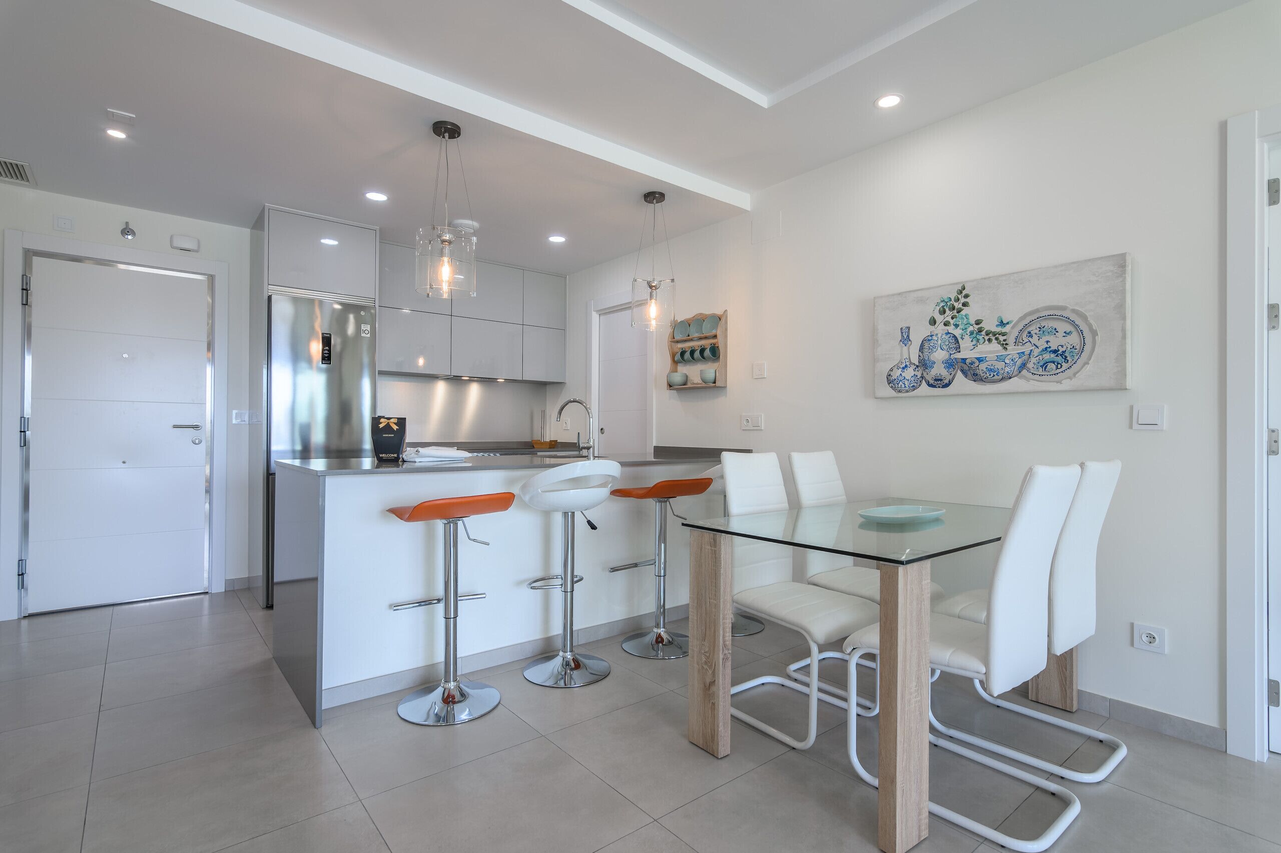 A modern kitchen with white cabinets, a stainless steel refrigerator, and a glass dining table with white chairs.