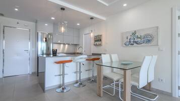 A modern kitchen with white cabinets, a stainless steel refrigerator, and a glass dining table with white chairs.