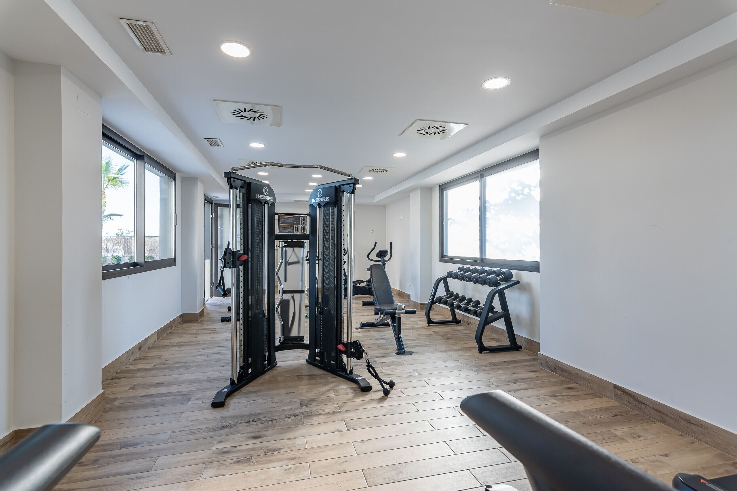 Un gimnasio moderno con piso de madera, equipamiento de ejercicio y grandes ventanales que permiten la entrada de luz natural.