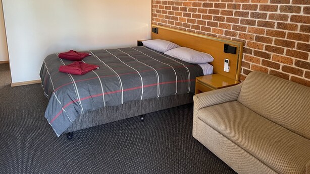 Habitación de hotel con cama, cabecera de madera, mesita de noche, termostato digital, pared de ladrillo y un cuadro en la pared.