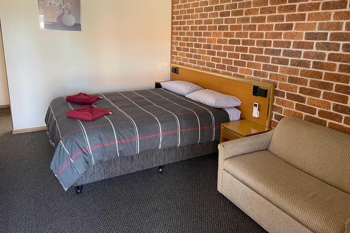 Ein Hotelzimmer mit einem Bett, einem Holz-Kopfende, einem Nachttisch, einem digitalen Thermostat, einer Ziegelwand und einem gerahmten Bild an der Wand.