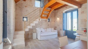 A rustic stone interior with a white sofa, wooden staircase, and a dining area. - Borgofreddo (Monopoli)