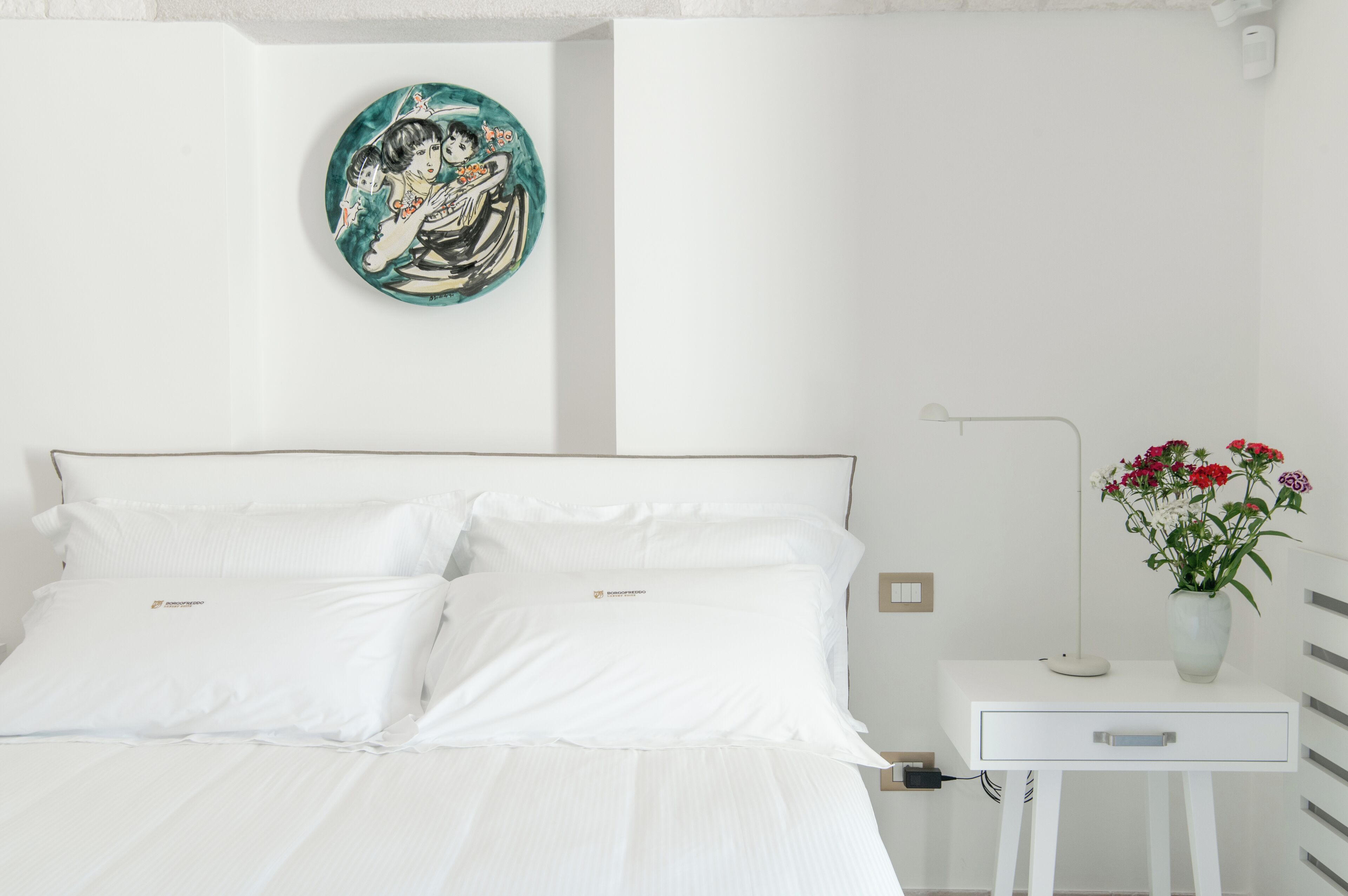 Una cama bien hecha con ropa blanca, una mesita de noche con un florero y un plato decorativo en la pared.