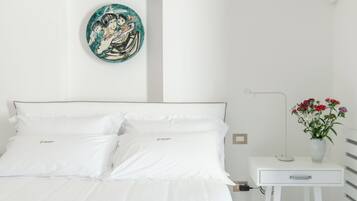 Una cama bien hecha con ropa blanca, una mesita de noche con un florero y un plato decorativo en la pared.