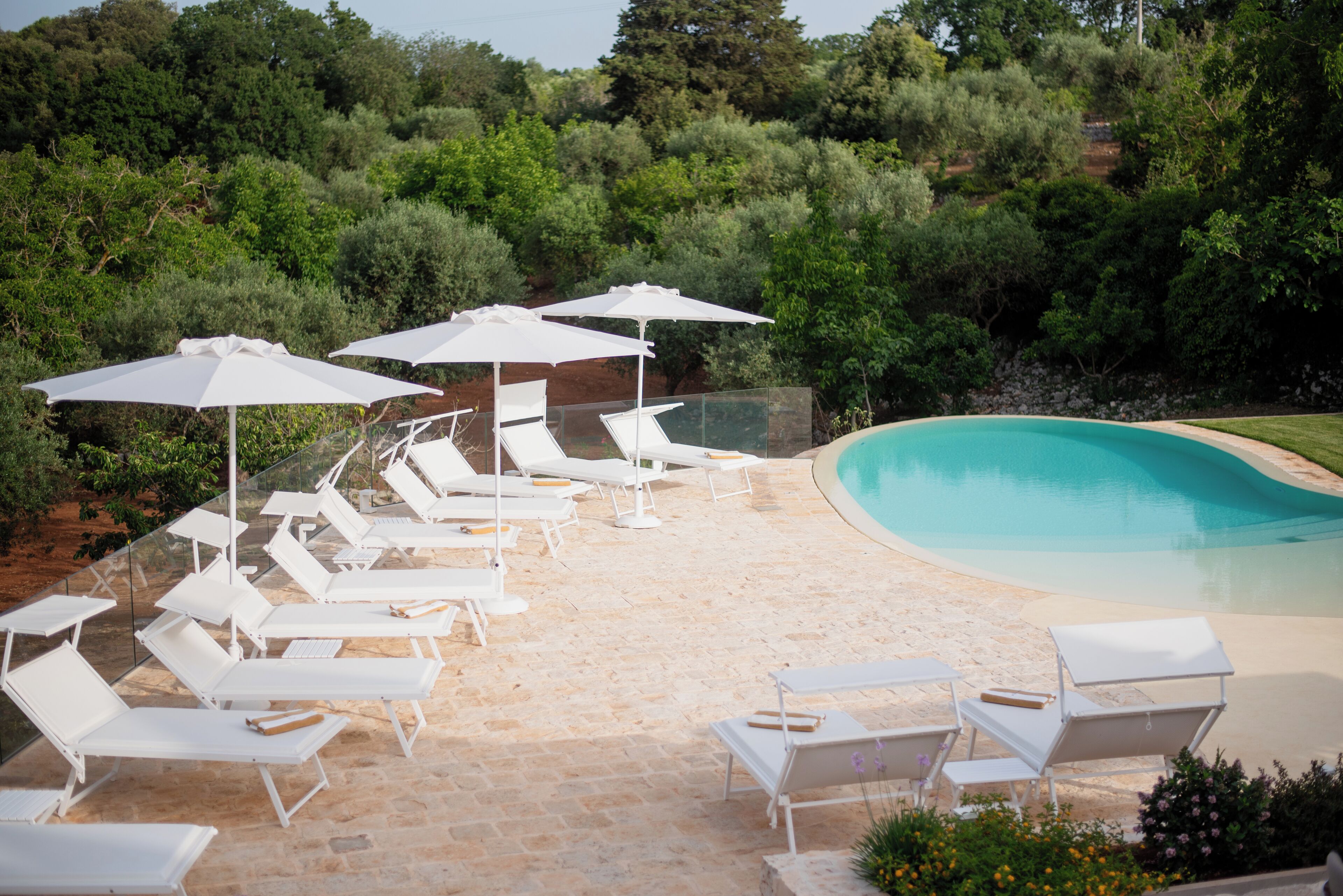 A poolside area with white lounge chairs and umbrellas, surrounded by greenery.