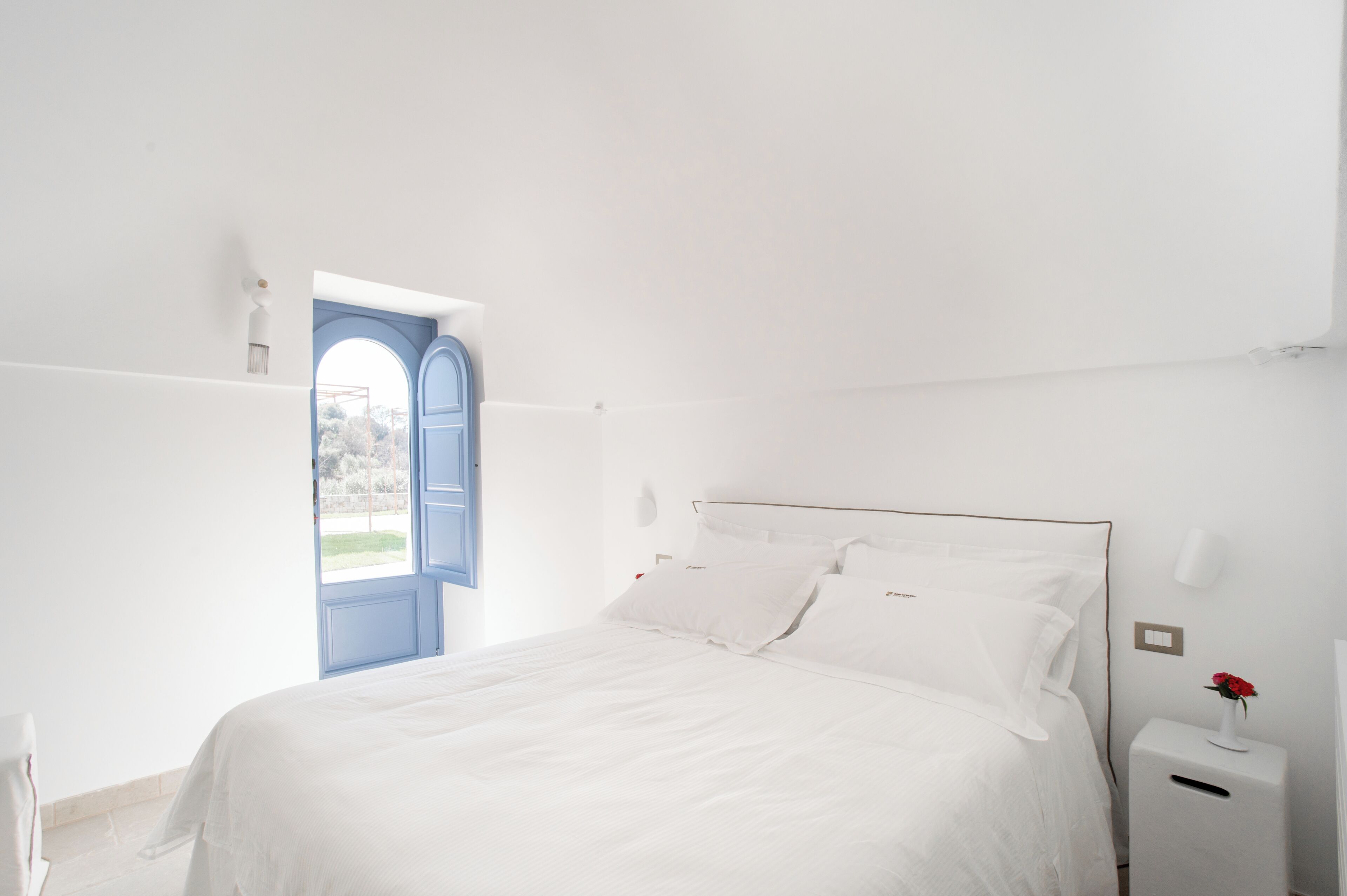 A white bedroom with a bed, a nightstand, a wall-mounted lamp, and a door leading to a balcony.