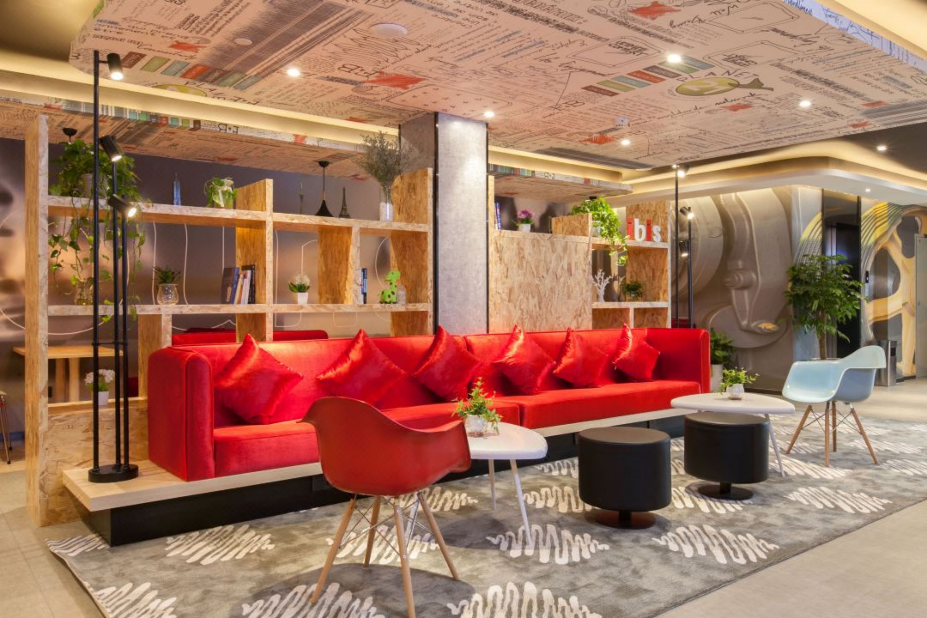 A modern interior with a red sofa, black ottoman, and a white table. There are shelves with plants and books, and a unique ceiling design.