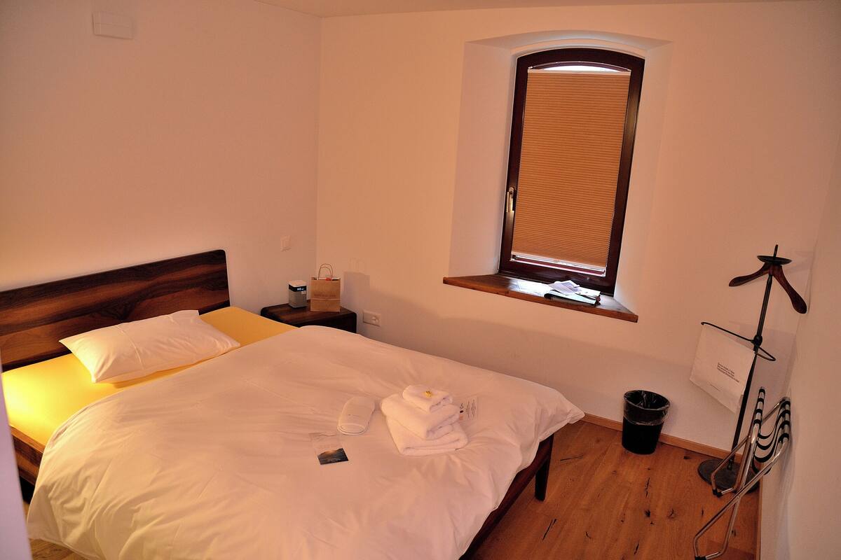 Cama bem arrumada com roupa branca, mesa de cabeceira com abajur, janela com persianas e uma lixeira.