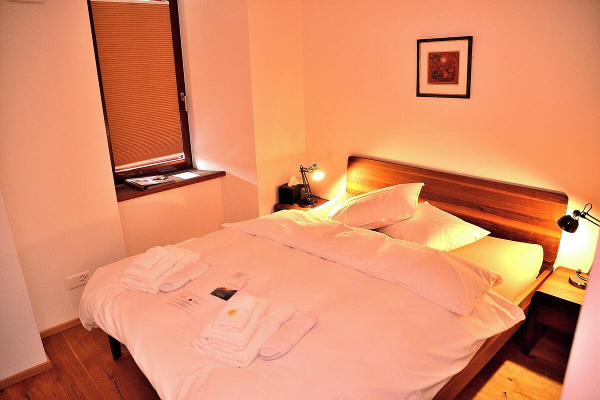 Uma cama bem arrumada com lençóis e toalhas brancos, uma luminária de cabeceira e um quadro na parede.