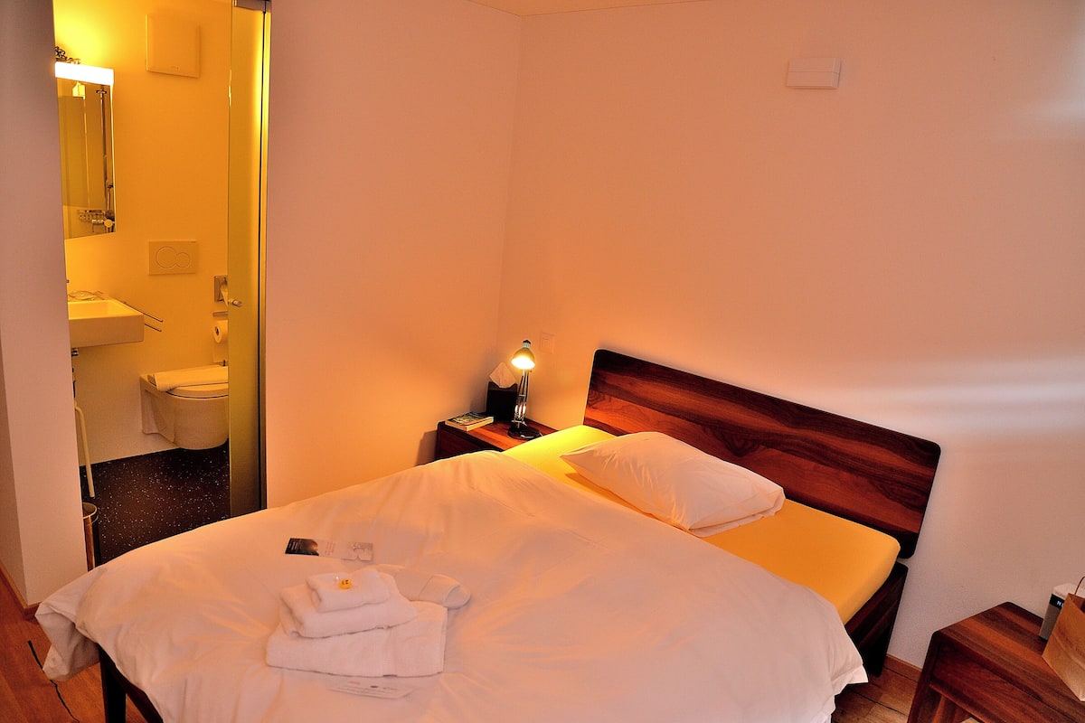 Quarto de hotel com uma cama, mesa de cabeceira com abajur, cabeceira de madeira e banheiro visível através de uma porta aberta.