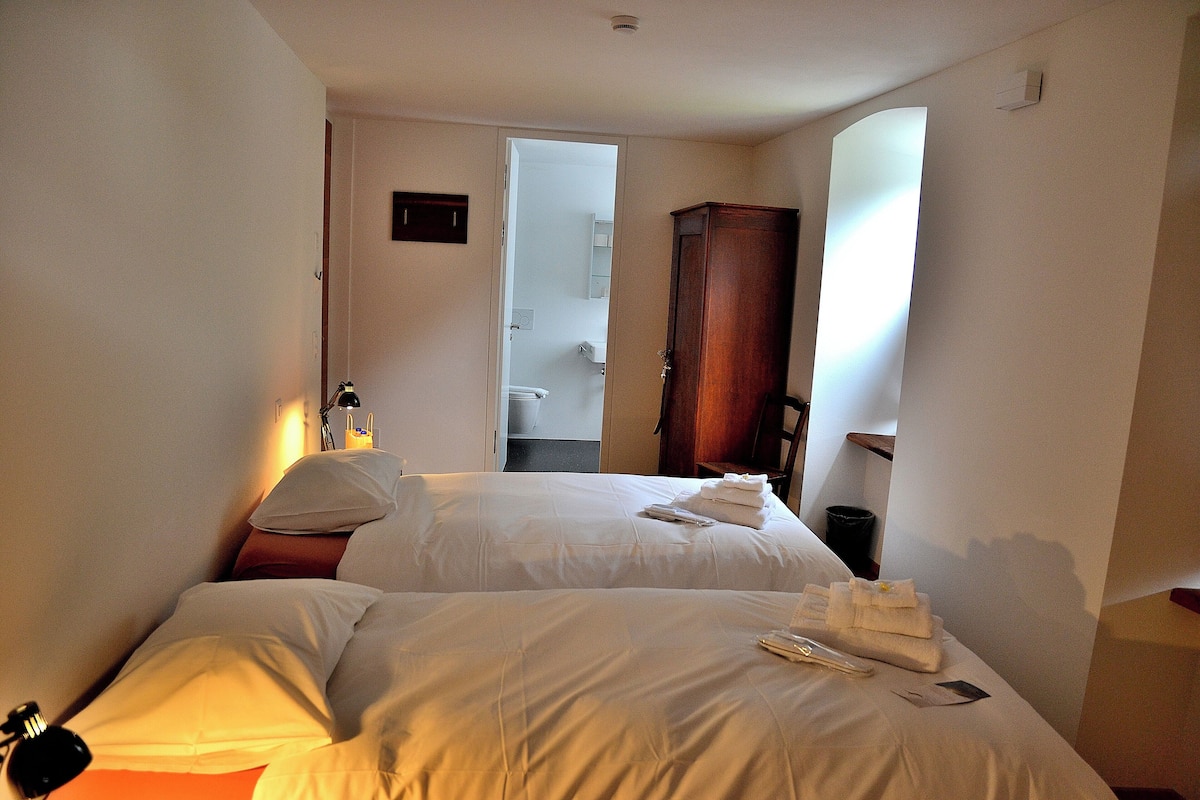 Quarto de hotel com duas camas, um guarda-roupa de madeira, um espelho e um banheiro visível por uma porta aberta.