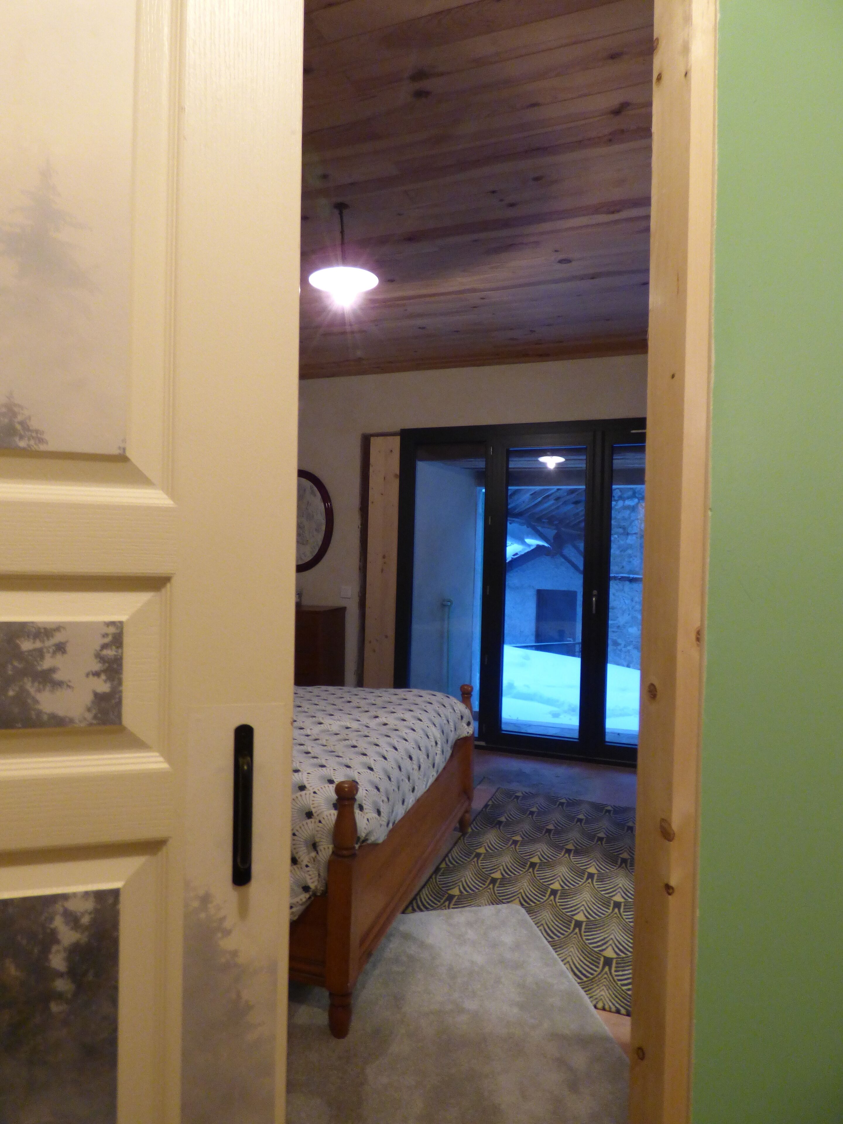 A bedroom with a wooden ceiling, a bed with a patterned bedspread, a door leading to a balcony with a view of the outside.