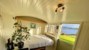 A cozy room with a bed, a potted plant, a window with a view, a small table with bottles, and a fire extinguisher. - A quiet and cosy Shepherds Hut, with wood burning fire with scenic views (Conwy)